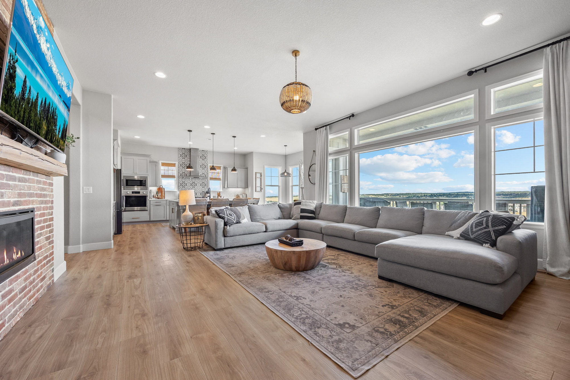 Open-concept living room with wide-plank floors, brick fireplace, large sectional sofa, and expansive windows overlooking the Front Range.